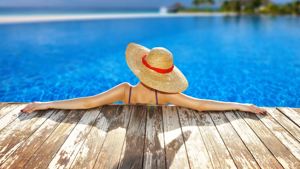 Ragazza prende il sole a bordo piscina con un cappello che la protegge dal sole - Come preparare la pelle al sole e proteggerla al meglio 